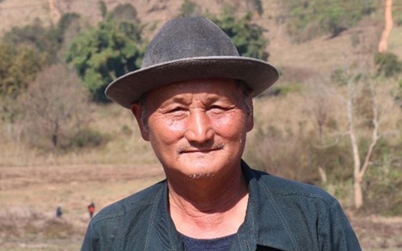 A man smiling at the camera, wearing a hat, in rural Laos.