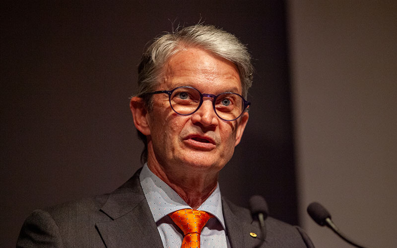 Professor Brendan Crabb, wearing an orange tie, speaking in front of podium microphones.