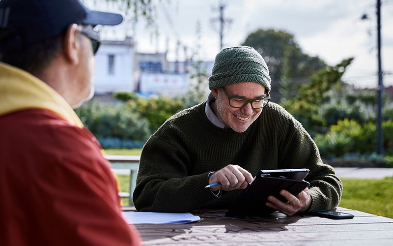 Peter Higgs, wearing a jumper and beanie on a park bench speaking to another person in the foreground.